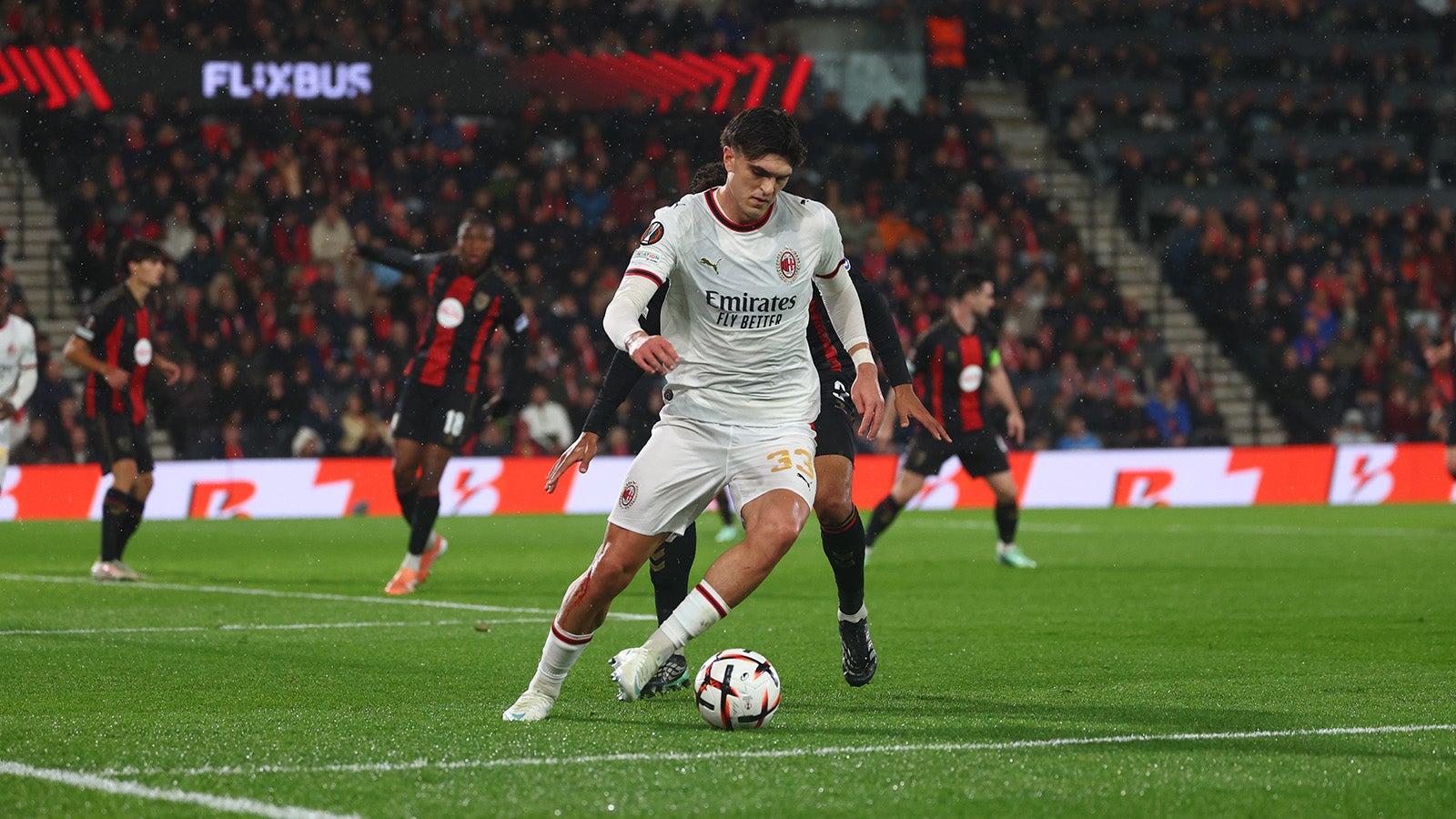 An AC Milan player in a white kit dribbles the ball in the rain during the Europa League match against Bournemouth