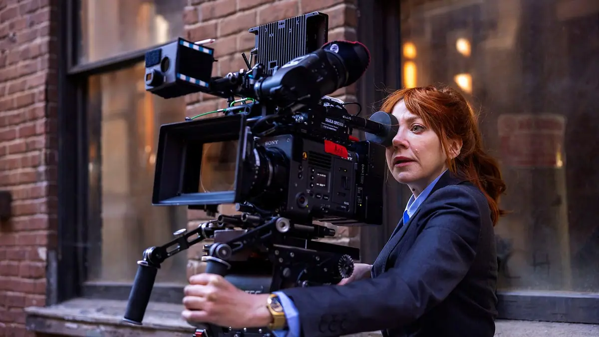 Diane Morgan as Philomena Cunk in a dark blazer, peering into a film camera on a rig outside a brick building