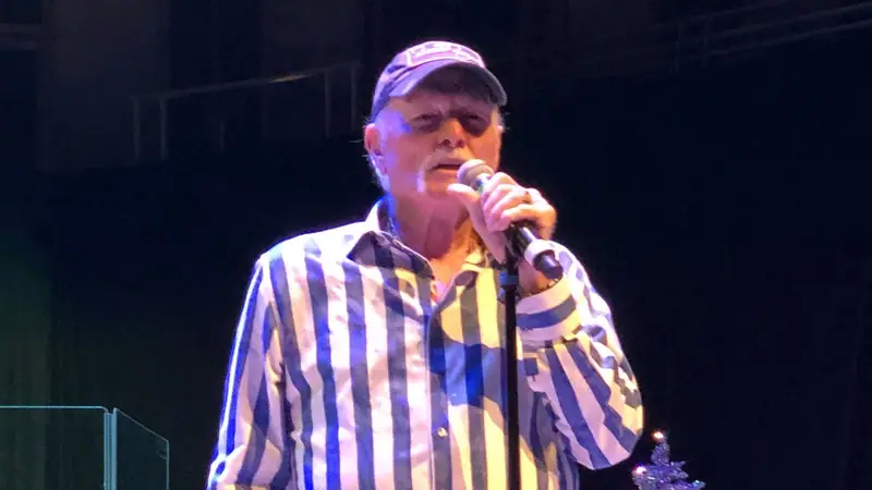 Mike Love of the Beach Boys singing into a microphone on stage in a striped shirt and cap