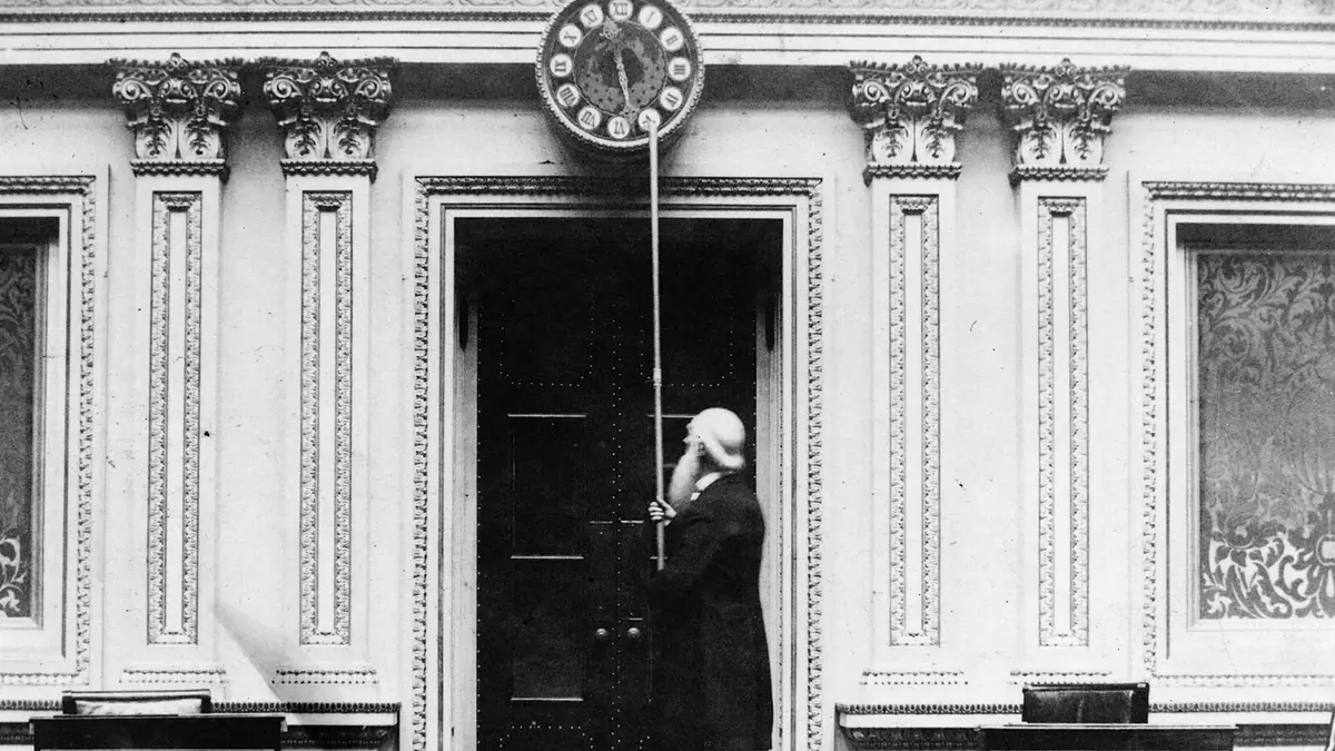Historic black-and-white photo of a man using a long pole to turn back the hands of a large wall clock in the U.S. Senate chamber